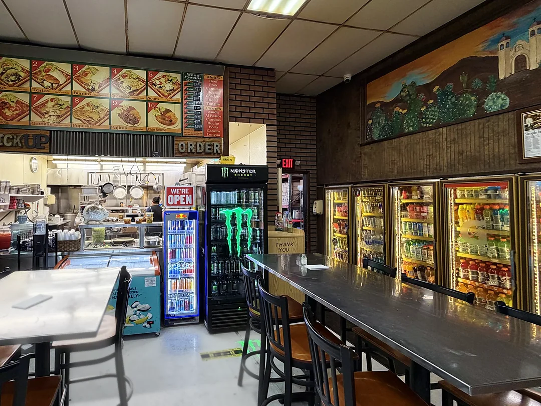 El comedor de la taquería: mesas de acero, la ventanilla de la cocina con el menú arriba y un mural de un pueblo mexicano en la pared